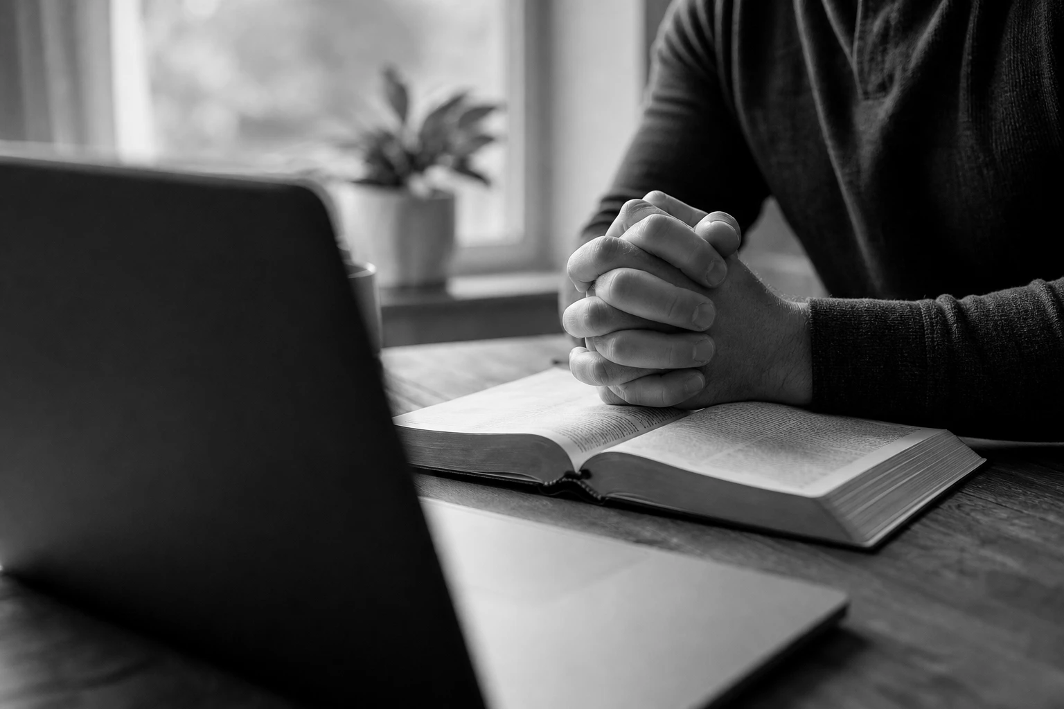 Hands folded in prayer over an open Bible at a laptop